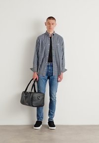 Melnas duffel soma ar diviem rokturiem, gludu tekstūru un pelēku krāsu. Modelis valkā svītraino krekliņu, melnu T-kreklu, zila džinsus un melnus sporta apavus.