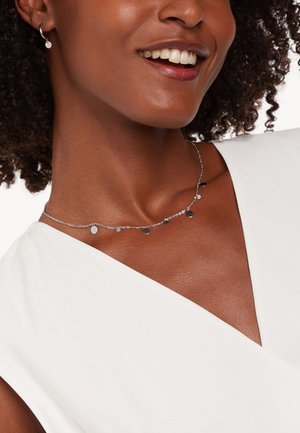 Zilveren ketting met cirkelvormige hangers van verschillende groottes, met een verfijnd schakeldesign. Gedeeld tegen een lichte stofachtergrond.