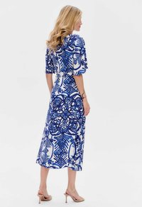 Niebieska i biała sukienka maxi w kwiatowy wzór, z krótkimi rękawami, zaznaczoną talią, lekkim materiałem i zwiewnym dołem. Widać sandały na wysokim obcasie.