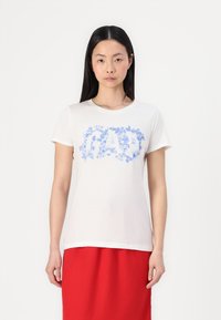 Weißes Baumwoll-T-Shirt mit kurzen Ärmeln, das vorne mit blauer Floralbeschriftung versehen ist. Das Model trägt einen roten Rock. Einfacher runder Ausschnitt.