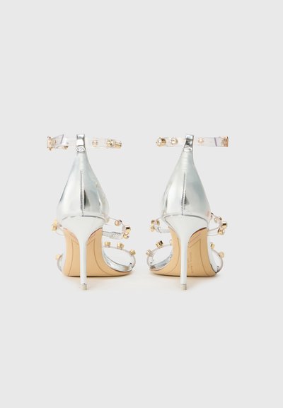 Sandale cu toc înalt, argintii, cu bretele transparente, având accente aurii și embellishments cu bijuterii. Design cu vârful ascuțit și toc stiletto.