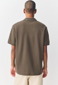 Hombre de pie, de espaldas, vistiendo una camiseta polo de mangas cortas color oliva y pantalones beige, frente a un fondo claro y liso.