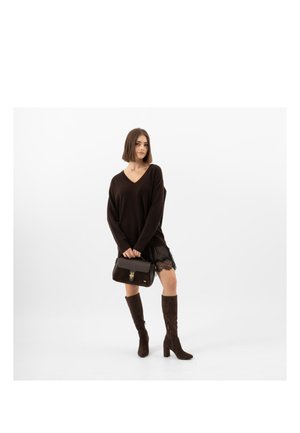 Femme portant un pull noir à col en V, une jupe bordée de dentelle, des bottes marron montant jusqu'au genou, tenant un sac à main marron foncé, posant devant un fond blanc uni.