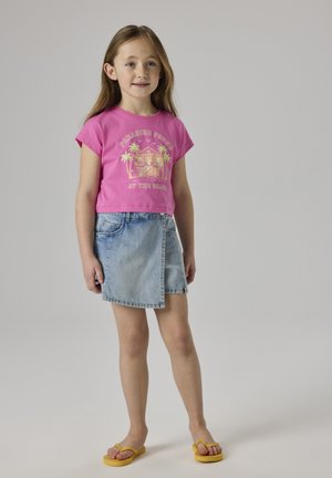 Niña joven con una camiseta gráfica rosa, falda cruzada de mezclilla y chanclas amarillas, de pie frente a un fondo claro y liso.
