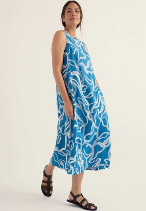 Žena v modrých šatách bez rukávov s bielymi abstraktnými vzormi a čiernymi plochými sandálmi so remienkami stojaca pred jednofarebným pozadím.