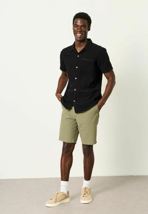 Mann steht lächelnd da, trägt ein schwarzes kurzärmeliges Hemd mit Knöpfen, olivgrüne Shorts, weiße Socken und beige Sneakers.