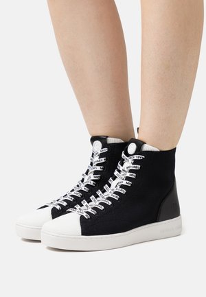 Des jambes portant des baskets montantes noir et blanc avec des semelles blanches et des lacets de marque "Michael Kors", se tenant sur un fond blanc.