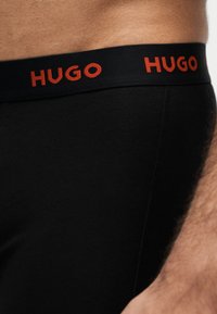 Gros plan d’une personne portant des sous-vêtements noirs avec une ceinture noire affichant le nom de la marque HUGO en lettres rouges.