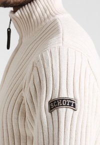 Krämfärgad ribbad stickad tröja med hög krage, försedd med en zippered hals och en svart broderad logotyp på axeln.