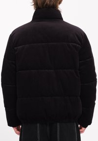 Schwarze Corduroy-Pufferjacke mit hohem Kragen, gestepptem Design, elastischem Bund und geripptem Material. Von hinten betrachtet.