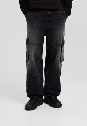 Person iført falmede sorte cargobukser med sidelommer og sorte sneakers, stående med hænderne i lommerne mod en grå baggrund.