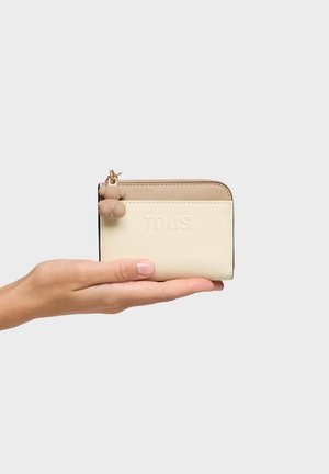 Mano che tiene un portafoglio Tous beige e crema con superficie texturizzata e piccolo ciondolo a forma di orsetto sulla cerniera, su sfondo bianco.