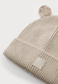 Bonnet beige en tricot avec un large revers côtelé, surface texturée et un petit haut arrondi. Présente une étiquette en tissu cousue sur le revers.