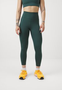 Ciemnozielone legginsy o wysokim stanie o gładkiej fakturze, połączone z jasn Yellowewymi butami sportowymi o fakturowanej podeszwie i sznurowadłach.
