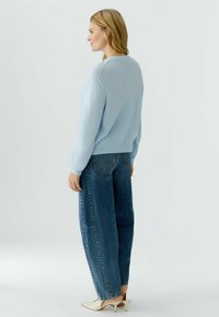 Frau steht seitlich, trägt einen hellblauen Pullover, lockere blaue Jeans und weiße Schuhe mit kleinem Absatz vor einem schlichten hellen Hintergrund.