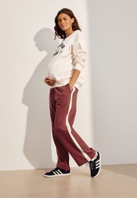 Hvid sweatshirt med trykt tekst, vinrøde joggingbukser med hvide sidelister og sorte sneakers med hvide detaljer. Model poserer med hånden på maven.