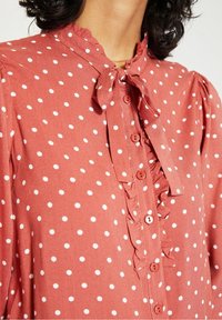 Rostfärgad blus med vita prickar, med volangdetaljer vid knapplisten, knäppning med knappar och en knuten halsringning. Korta, rynkade ärmar.