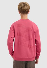 Jongere met kort blond haar, gekleed in een roze sweatshirt met reliëf zigzagletters op de achterkant, gecombineerd met een bruine broek.
