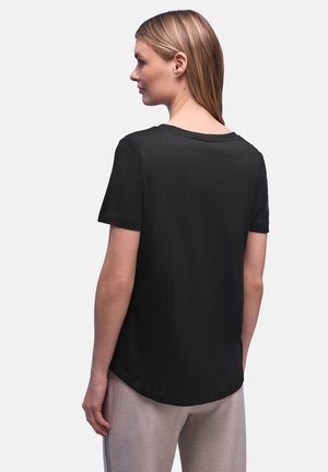 Kobieta o jasnobrązowych włosach ubrana w prostą czarną koszulkę z krótkim rękawem i jasne spodnie, zwrócona tyłem na jednolitym tle.