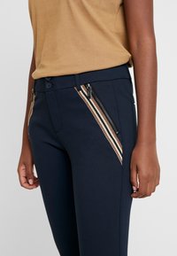 Marina byxor med kontrasterande randiga sidopaneler och fickor med dragkedja. Tillverkade av slätt tyg med en figurnära design och knäppning.