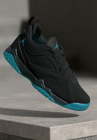 Sneaker atletica nera e teal con dettagli in rete e pelle, sospeso sopra una superficie di pietra, evidenziando la suola e i lacci.