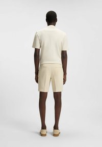 Camicia corta a maniche corte color crema abbinata a pantaloni corti beige chiaro. Il tessuto appare liscio con una vestibilità regolare. Le scarpe sono beige con una suola in legno.