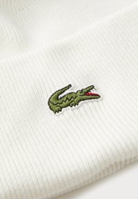 Tecido alb cu nervuri, cu un mic logo brodat reprezentând un crocodil verde cu gura deschisă roșie.