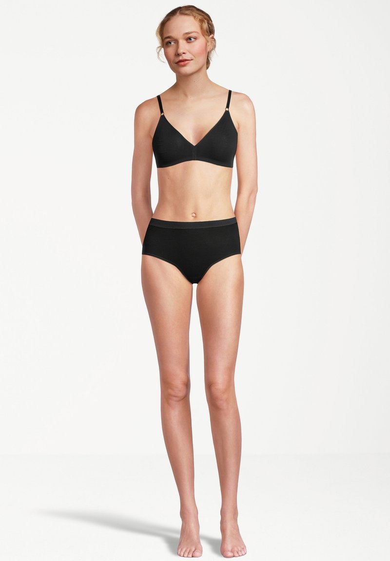 Donna in piedi a piedi nudi, indossando un reggiseno nero e slip a vita alta, contro uno sfondo bianco semplice, che guarda leggermente di lato.