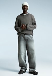 Grå ribbet sweater, løse grå denim bukser med falming, og mørke sneakers. Modellen bærer en hvid strikket beanie og en sølv kædede halskæde.