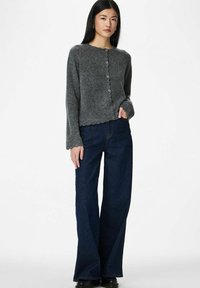 Pelēks adīts kardigans ar viļņotu apakšu un pogām, kombinēts ar vaļīgi pieguļošām tumši zilām džinsa biksēm. Modelis stāv ar rokām kabatās.