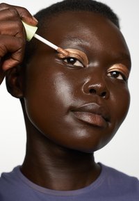 Applicateur d'ombre à paupières liquide dans la main, appliquant une teinte dorée scintillante sur la paupière. Peau foncée avec une texture lisse, portant une chemise violette.