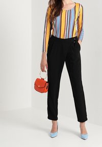 Långärmad randig topp i blått, gult och orange. Svarta skräddarsydda byxor med en subtil lyster. Håller en liten röd handväska med ett runt handtag.