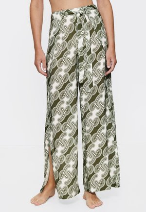 Woman wearing high-waisted wide-leg pants with green and white abstract pattern and side slits, barefoot against a white background.