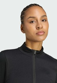 Black zip-up sports top with high collar, textured fabric, and subtle logo detail on the chest. Smooth finish and fitted design.