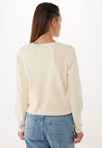 Kvinna med långt brunt hår som bär guldörhängen, en off-white långärmad tröja och ljusblå denimjeans, vänd bort mot en vit bakgrund.