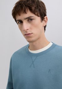 Joven con cabello corto castaño, vistiendo un suéter azul de cuello redondo sobre una camisa blanca, mirando hacia adelante con una expresión neutral.