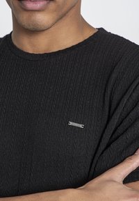 Czarny sweter z teksturowanego dzianiny o prążkowanym wzorze, z metalową metką marki "Kavanagh" na lewej stronie klatki piersiowej. Okrągły dekolt.