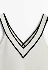 Top bianco senza maniche con profondo scollo a V, bordato da strisce nere a contrasto. Il tessuto ha una texture a coste.