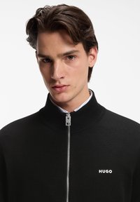 Νεαρός άνδρας με καστανά μαλλιά που φοράει μαύρο πουλόβερ με φερμουάρ και το λογότυπο "HUGO" στο στήθος, μπροστά σε λευκό φόντο.