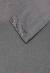 Pelēks adīts džemperis ar ribotām piedurknēm. Tuvplāns, kas parāda smalko tekstūru un vienmērīgu šuvju kvalitāti. Vienkāršs, bezšuvju dizains.