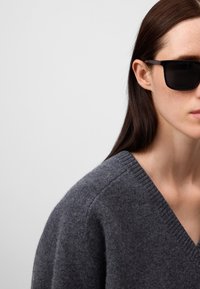 Donkergrijs wollen sweater met een V-halsontwerp, gedragen met zwarte rechthoekige zonnebril. Glad textuur met een zachte, gezellige uitstraling.