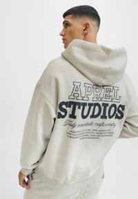 Šedá mikina s kapucí vyrobená z měkké tkaniny, s výrazným černým potiskem na zadní straně, který obsahuje "APREL STUDIOS" a popisný text.