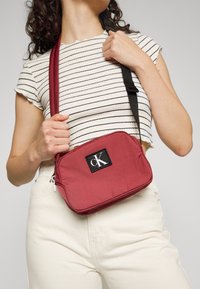 Borsa a tracolla Calvin Klein in tessuto rosso, forma rettangolare, con una patch logo nera, chiusura con zip e tracolla regolabile.
