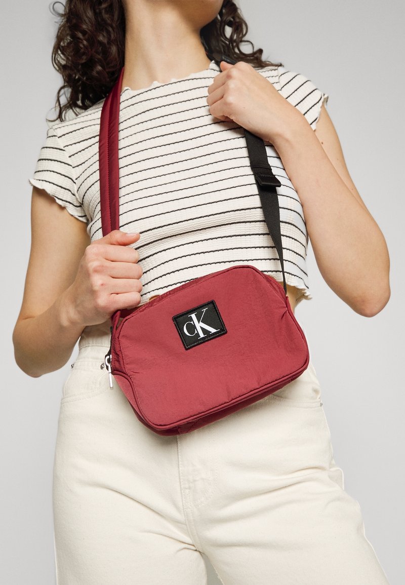 Borsa a tracolla Calvin Klein in tessuto rosso, forma rettangolare, con una patch logo nera, chiusura con zip e tracolla regolabile.