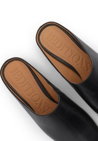 Chaussures en cuir noir à enfiler avec une texture lisse, présentant une semelle intérieure beige embossée du mot "EDITIONS" et un design à dos ouvert.