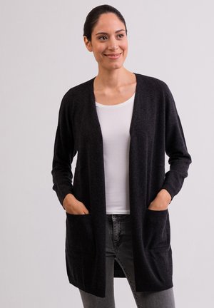 Cardigan grigio scuro in morbido tessuto, design aperto davanti, polsini a costine e due tasche frontali. Il modello indossa un top bianco e jeans grigi.