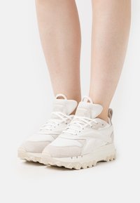 Valkoiset ja beige-lace-up paksupohjaiset lenkkikengät paljailla jaloilla, nähtynä edestä valkoista taustaa vasten.