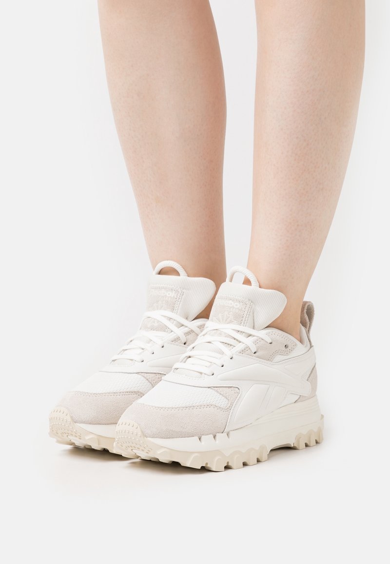 Valkoiset ja beige-lace-up paksupohjaiset lenkkikengät paljailla jaloilla, nähtynä edestä valkoista taustaa vasten.