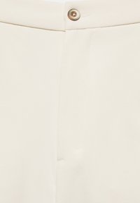Gros plan sur la ceinture d’un pantalon crème avec une fermeture à bouton et une fermeture éclair dissimulée, montrant un tissu lisse et une coupe épurée.