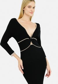 La modella posa in un vestito nero attillato con scollatura a V profonda e un dettaglio intrecciato in vita, con bordi beige a contrasto sulle maniche.